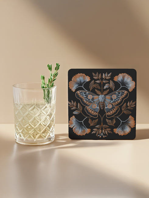Square black ceramic coaster with detailed copper and blue moth illustration centered among symmetrical botanical elements including ginkgo-like leaves and delicate foliage.